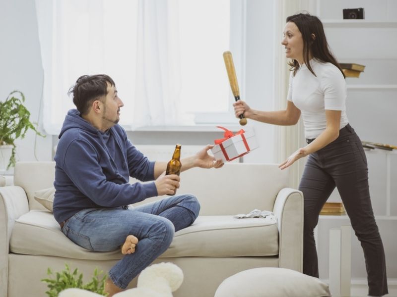 A husband is drinking beer while her wife beats him with a bat