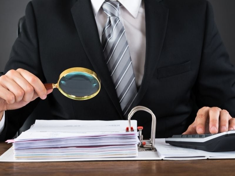 Businessman Inspecting Invoice With Magnifying Glass At Wooden Desk
